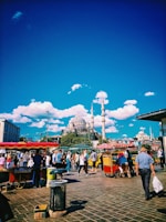 A bustling street scene with a lively crowd walking past food stalls under a clear blue sky. A prominent mosque with multiple domes and tall minarets is visible in the background, surrounded by lush green trees. The image also includes vibrant red and yellow colors from the food carts, creating a dynamic atmosphere.