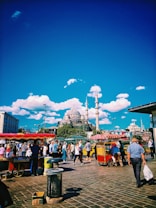 A bustling street scene with a lively crowd walking past food stalls under a clear blue sky. A prominent mosque with multiple domes and tall minarets is visible in the background, surrounded by lush green trees. The image also includes vibrant red and yellow colors from the food carts, creating a dynamic atmosphere.