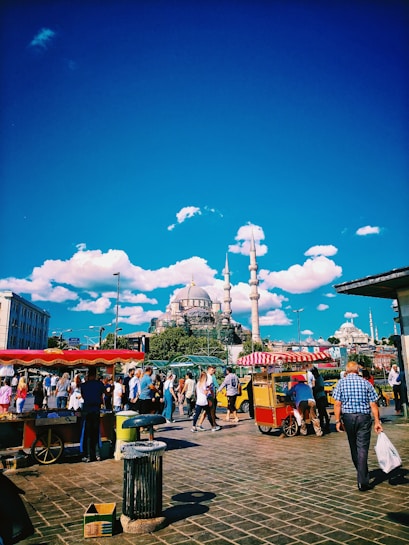 A bustling street scene with a lively crowd walking past food stalls under a clear blue sky. A prominent mosque with multiple domes and tall minarets is visible in the background, surrounded by lush green trees. The image also includes vibrant red and yellow colors from the food carts, creating a dynamic atmosphere.