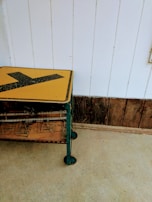 A vintage wire rack with a yellow and black directional sign on top is placed against a white paneled wall. The rack has weathered metal surfaces and a wooden shelf. The floor is concrete, and the lower part of the wall features rustic wooden planks.