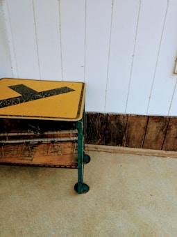 A vintage wire rack with a yellow and black directional sign on top is placed against a white paneled wall. The rack has weathered metal surfaces and a wooden shelf. The floor is concrete, and the lower part of the wall features rustic wooden planks.