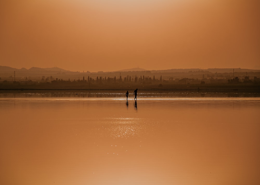 silhouette of 2 people standing on body of water during sunset