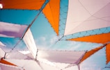 Geometric shapes are formed by interconnected white and orange fabric panels, set against a clear blue sky. The panels are structured with black netting and borders, creating an abstract and artistic canopy.