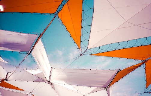 Geometric shapes are formed by interconnected white and orange fabric panels, set against a clear blue sky. The panels are structured with black netting and borders, creating an abstract and artistic canopy.