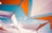Geometric shapes are formed by interconnected white and orange fabric panels, set against a clear blue sky. The panels are structured with black netting and borders, creating an abstract and artistic canopy.