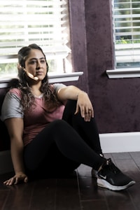 A woman sits on a dark wooden floor next to a wall with purple wallpaper, under partially opened blinds letting in strips of light. She wears a pink and gray shirt with black leggings and sneakers, with an expression suggesting deep thought.