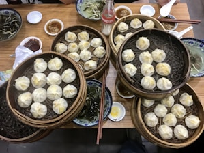 A wooden table is filled with several bamboo steamers containing steamed dumplings. There are blue and white plates with soup, small bowls with condiments like soy sauce and chili paste, and a glass bottle of beverage on the table. Chopsticks and ceramic spoons are also present.