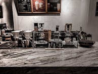 An array of coffee brewing equipment is displayed on a marble countertop. There are several different types of coffee makers, including pour-over and siphon coffee makers, as well as containers and jars filled with coffee beans. Behind the counter, decorative artwork featuring abstract patterns and figures adorns the wall. The ambiance is enhanced by low, warm lighting.