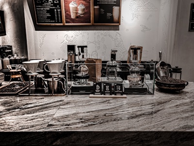 An array of coffee brewing equipment is displayed on a marble countertop. There are several different types of coffee makers, including pour-over and siphon coffee makers, as well as containers and jars filled with coffee beans. Behind the counter, decorative artwork featuring abstract patterns and figures adorns the wall. The ambiance is enhanced by low, warm lighting.