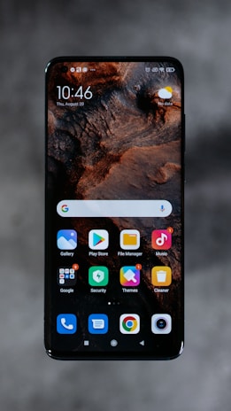 A smartphone displaying a home screen with various app icons, including Gallery, Play Store, File Manager, and Music. The background features a textured, earthy design. The time reads 10:46 and the date is displayed as Thursday, August 20. The top right corner shows a weather widget with a sun icon and a 'No data' label.