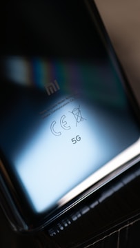 A close-up view of a smartphone's reflective surface, displaying the Xiaomi logo, CE certification, and 5G capabilities with a soft gradient of blue and black tones creating a sleek and modern appearance.