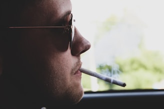A person wearing sunglasses is seen in profile, holding a lit cigarette in their mouth. Light smoke drifts upward against a blurred outdoor background.