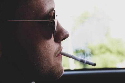 A person wearing sunglasses is seen in profile, holding a lit cigarette in their mouth. Light smoke drifts upward against a blurred outdoor background.