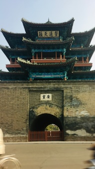 A traditional Chinese architectural structure with multiple tiers, featuring intricate wooden details and decorative elements. The building has a prominent arched entrance at its base, adorned with Chinese characters. The structure is characterized by its use of dark wood, green, and blue accents, and stands atop a stone base.