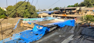 A series of rooftops covered with various tarps, predominantly blue and gray, appear in the foreground. The structures are older and somewhat dilapidated, with visible signs of wear and tear. In the background, there are several apartment buildings with balconies and more trees, creating an urban landscape. Electrical or telephone wires are visible crossing the scene.