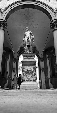 A classical statue is placed prominently at the center of an architectural setting, surrounded by ornate columns and arches. The statue stands on a detailed pedestal featuring decorative carvings. Two people are visible in the background, observing the artwork.