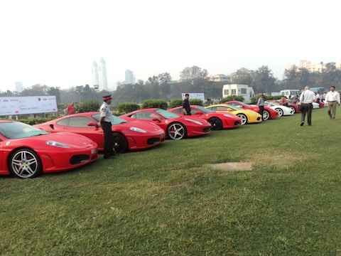 A lineup of luxury sports cars is parked on a grassy field with several people standing nearby. The cars, primarily red, include models with sleek designs and distinctive hubcaps. A uniformed security guard stands next to one of the cars, while other individuals appear to be observing or inspecting the vehicles. In the background, tall buildings and trees can be seen, along with banners indicating a car show.