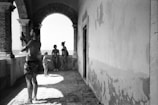 Four people, two adults and two children, are captured in a black-and-white photograph on a coastal terrace. One adult is taking a photo with a smartphone, while the others are standing near an archway, looking out toward the sea. The architecture is characterized by brick arches and worn walls, suggesting an aged or rustic setting.