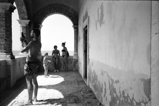 Four people, two adults and two children, are captured in a black-and-white photograph on a coastal terrace. One adult is taking a photo with a smartphone, while the others are standing near an archway, looking out toward the sea. The architecture is characterized by brick arches and worn walls, suggesting an aged or rustic setting.