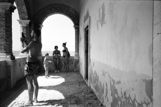 Four people, two adults and two children, are captured in a black-and-white photograph on a coastal terrace. One adult is taking a photo with a smartphone, while the others are standing near an archway, looking out toward the sea. The architecture is characterized by brick arches and worn walls, suggesting an aged or rustic setting.