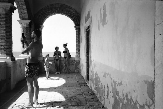 Four people, two adults and two children, are captured in a black-and-white photograph on a coastal terrace. One adult is taking a photo with a smartphone, while the others are standing near an archway, looking out toward the sea. The architecture is characterized by brick arches and worn walls, suggesting an aged or rustic setting.