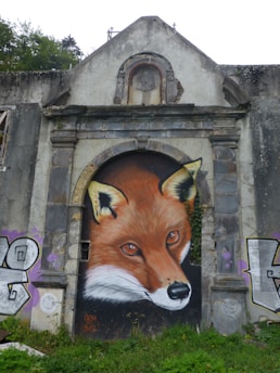 A large and detailed mural of a fox's face is painted over an old, stone archway. The naturalistic artwork highlights the fox's intense eyes and vibrant fur. Surrounding the mural, the building appears weathered, with some graffiti on either side and greenery at the base, suggesting an urban and possibly abandoned environment.