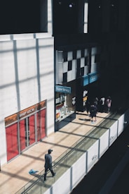 A shopping mall interior with a long hallway lined with shops. Sunlight casts geometric shadows on the walls and floor. One person is walking along a glass-railed walkway, and groups of people stand near shop entrances featuring signs like 'Shop Local' and 'Spring.'