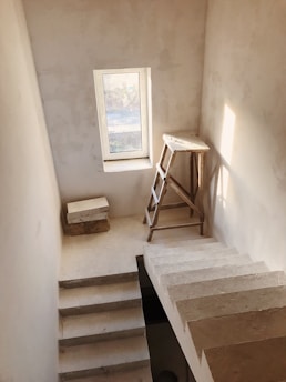 A minimalistic interior staircase with an unfinished look. The staircase is made of concrete or stone, and there is a wooden ladder leaning against one wall. A small window allows natural light to flood into the space, casting soft shadows on the walls.