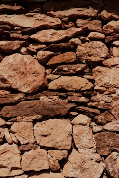 A rustic stone wall is constructed with irregularly shaped and sized rocks, tightly fitted together. The stones exhibit a range of earthy tones, with visible textures and weathering marks, suggesting an aged structure. Light and shadow interplay across the surface, highlighting the rough and uneven surface of the stones.
