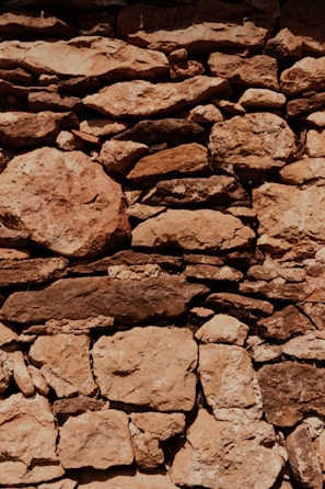 A rustic stone wall is constructed with irregularly shaped and sized rocks, tightly fitted together. The stones exhibit a range of earthy tones, with visible textures and weathering marks, suggesting an aged structure. Light and shadow interplay across the surface, highlighting the rough and uneven surface of the stones.