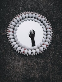 A circular mirror surrounded by an ornate frame made of seashells and small red and black beads. The reflection in the mirror shows an upright human hand against a dark background, creating a mysterious and intriguing visual effect.