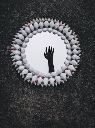 A circular mirror surrounded by an ornate frame made of seashells and small red and black beads. The reflection in the mirror shows an upright human hand against a dark background, creating a mysterious and intriguing visual effect.