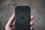 A hand holding a rugged black smartphone with a textured back cover and the CAT logo prominently displayed. The camera and flash can be seen on the phone's back in a vertical alignment.