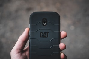 A hand holding a rugged black smartphone with a textured back cover and the CAT logo prominently displayed. The camera and flash can be seen on the phone's back in a vertical alignment.