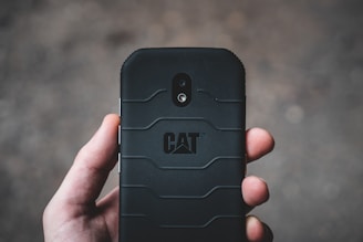 A hand holding a rugged black smartphone with a textured back cover and the CAT logo prominently displayed. The camera and flash can be seen on the phone's back in a vertical alignment.