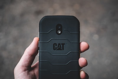 A hand holding a rugged black smartphone with a textured back cover and the CAT logo prominently displayed. The camera and flash can be seen on the phone's back in a vertical alignment.