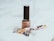 A beige nail polish bottle with a black cap is placed on a soft white fabric. Nearby, there is an ornate fountain pen with floral designs and gold accents. The scene suggests a subtle elegance and minimalistic aesthetics.
