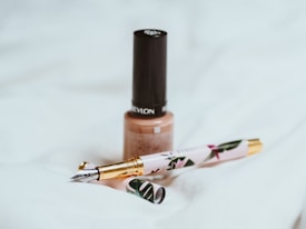 A beige nail polish bottle with a black cap is placed on a soft white fabric. Nearby, there is an ornate fountain pen with floral designs and gold accents. The scene suggests a subtle elegance and minimalistic aesthetics.