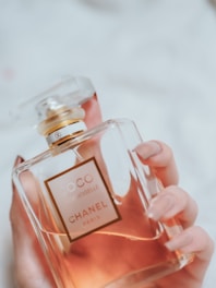 A hand with manicured nails holds a glass bottle of Coco Mademoiselle Chanel perfume. The bottle has a transparent cap and a golden neck. The liquid inside appears amber or rosy in color.