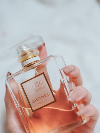 A hand with manicured nails holds a glass bottle of Coco Mademoiselle Chanel perfume. The bottle has a transparent cap and a golden neck. The liquid inside appears amber or rosy in color.