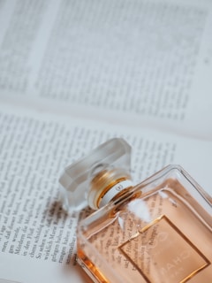 A classic perfume bottle lies on an open book. The bottle is rectangular with a clear stopper, and the liquid inside is a warm amber color. The book pages are filled with printed text, providing a soft background.