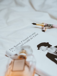 A soft focus image featuring a book titled 'Die Kunst, Chanel zu sein' next to a sophisticated perfume bottle with reflections of ambient light. Beside the book lies an elegant fountain pen with gold accents, resting on a background of white fabric.