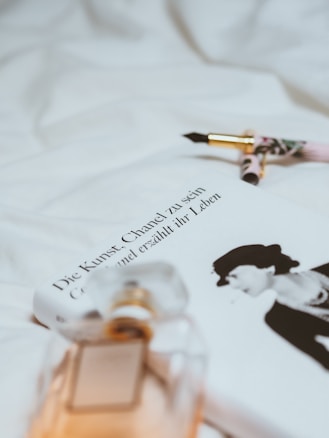 A soft focus image featuring a book titled 'Die Kunst, Chanel zu sein' next to a sophisticated perfume bottle with reflections of ambient light. Beside the book lies an elegant fountain pen with gold accents, resting on a background of white fabric.