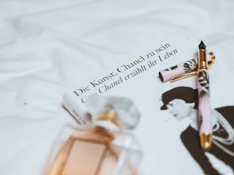 A book with the title 'Die Kunst - Chanel zu sein' is placed on a white fabric surface. Next to it, a decorative pen with floral patterns and gold accents rests atop the book. In the foreground, there is a blurred image of a perfume bottle with a geometric design, and the book cover features a black and white photograph of a woman.