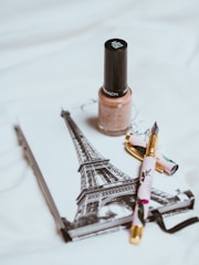 A bottle of beige nail polish is placed on top of a notebook featuring a black and white image of the Eiffel Tower. Nearby lays a decorative pen with floral patterns, featuring a gold nib.