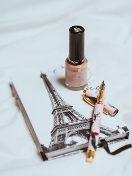 A bottle of beige nail polish is placed on top of a notebook featuring a black and white image of the Eiffel Tower. Nearby lays a decorative pen with floral patterns, featuring a gold nib.