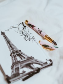 A decorative pen rests on a notebook featuring a monochromatic illustration of the Eiffel Tower and bare tree branches. The pen has a floral design and gold accents.