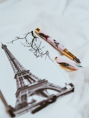 A decorative pen rests on a notebook featuring a monochromatic illustration of the Eiffel Tower and bare tree branches. The pen has a floral design and gold accents.