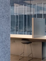 A modern office setup featuring a sleek stool placed under a light wooden desk. The backdrop includes large glass windows that reveal a view of an adjacent building's architecture with vertical lines. The interior design incorporates minimalist elements with a focus on clean lines and open space.