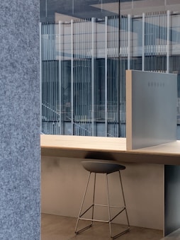 A modern office setup featuring a sleek stool placed under a light wooden desk. The backdrop includes large glass windows that reveal a view of an adjacent building's architecture with vertical lines. The interior design incorporates minimalist elements with a focus on clean lines and open space.